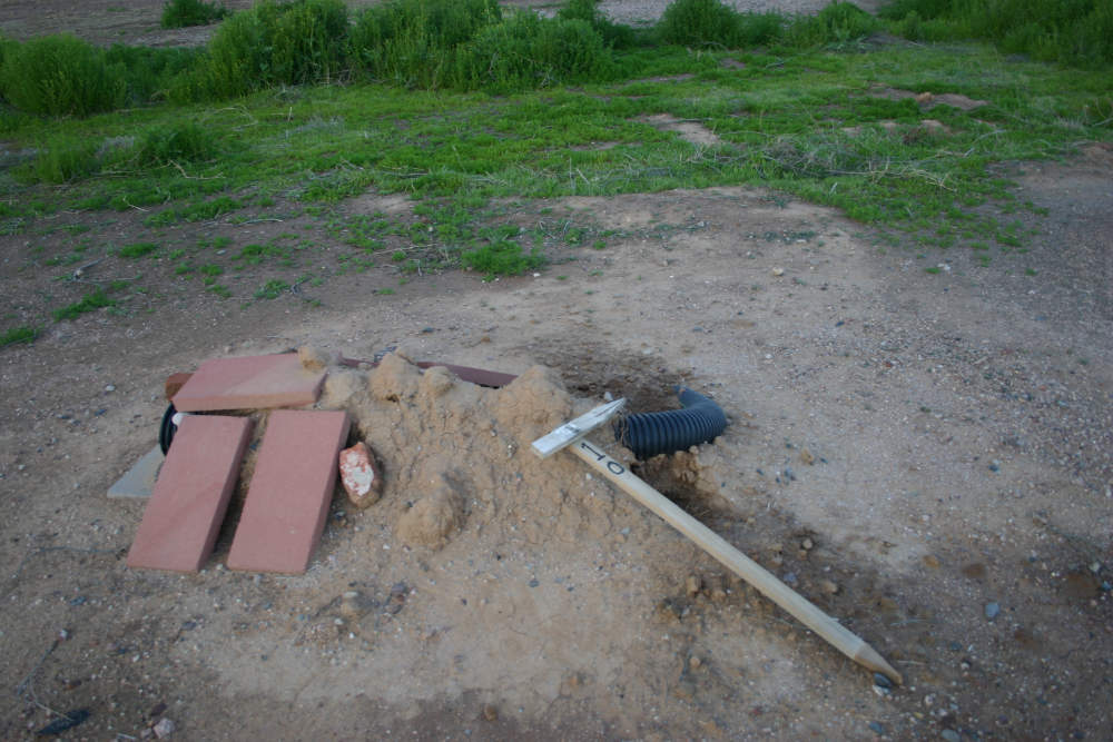 An abandoned artificial burrow at the ASU Polytechnic campus. This burrow's weeping tile has been uncovered as it leads down to the main burrow area. There are large narrow red bricks covering the entrance and a grey cinder block under the entrance. A wooden perch has fallen over on top of the burrow. 