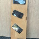 Four cellphones with broken screens are attached to a wooden board.