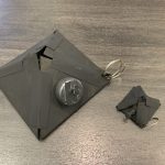 A silver pendant sits on a grey, cardboard pyramid. A smaller one sits next to it.