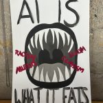 A poster displays the phrase: "AI IS WHAT IT EATS," with an image of a monstrous mouth eating words like racism, ableism, sexism, and classism.