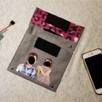 A fabric pouch lies next to a phone. The front of the pouch has realistic photos of children with black circles covering their faces.